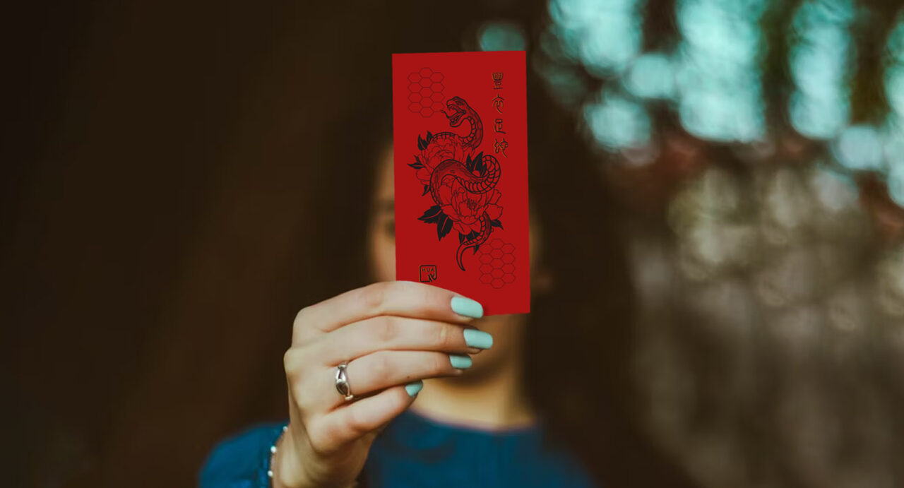 A person holding a Lunar New Year red envelope adorned with a detailed illustration of a snake surrounded by floral designs, against a blurred green and brown background. The person's face is out of focus, with attention drawn to the envelope and their teal-painted nails.